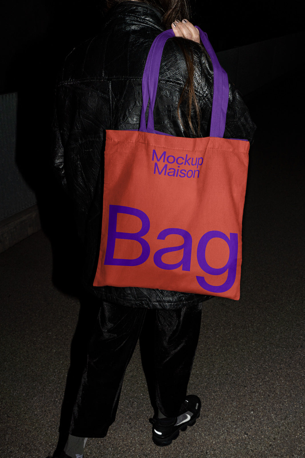 PSD mockup of an orange tote bag with purple text, held by a person in a dark outfit. The bag reads "Mockup Maison Bag."