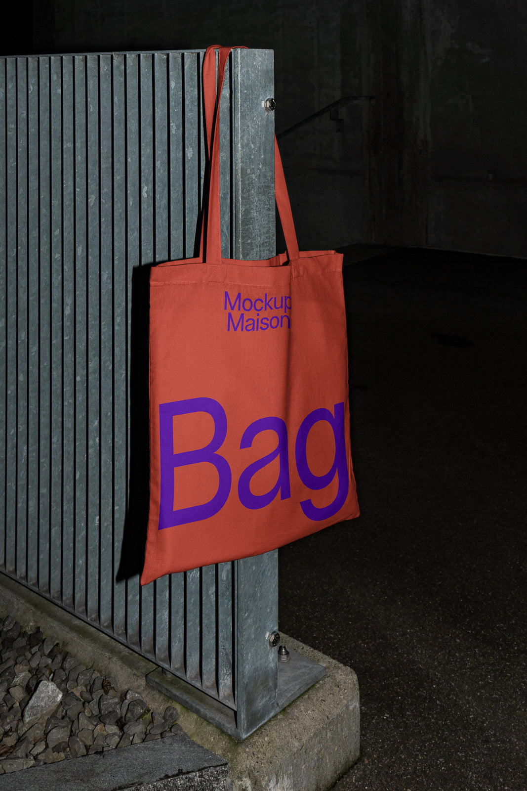 PSD mockup of a red tote bag with purple text hanging on a metal fence, displaying the words "Mockup Maison" and "Bag" in a dimly lit setting.