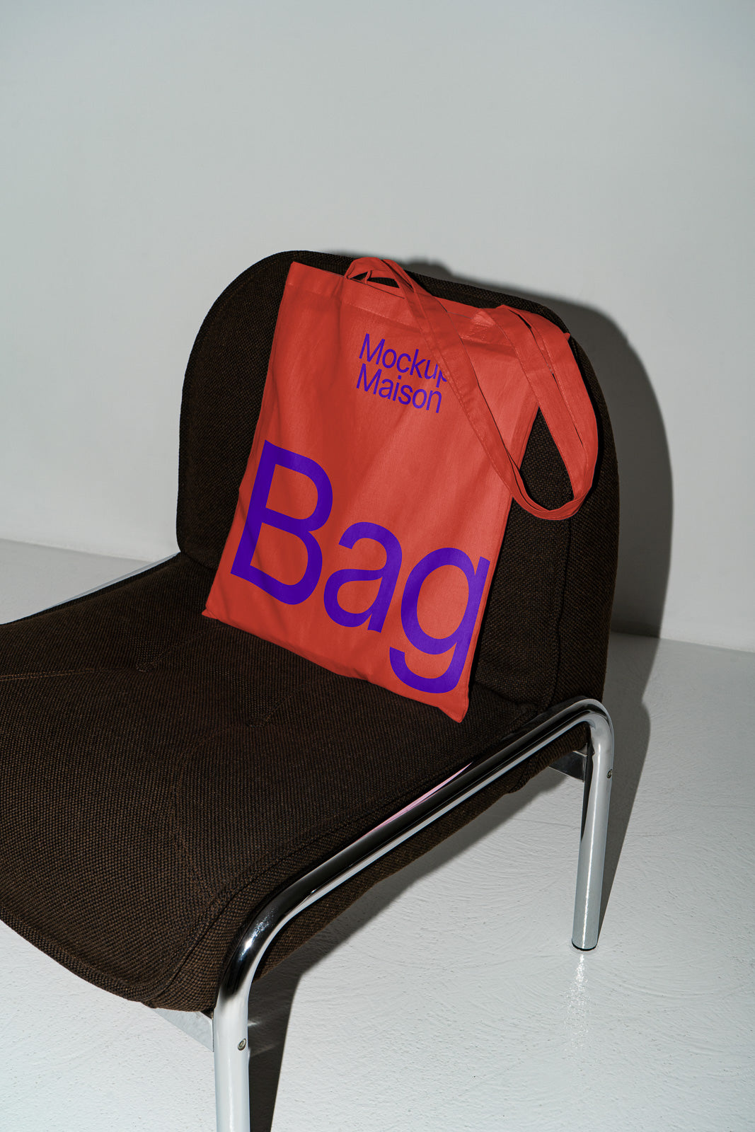 PSD mockup of a red tote bag with "Mockup Maison" and "Bag" in blue text, placed on a dark brown chair with chrome legs.