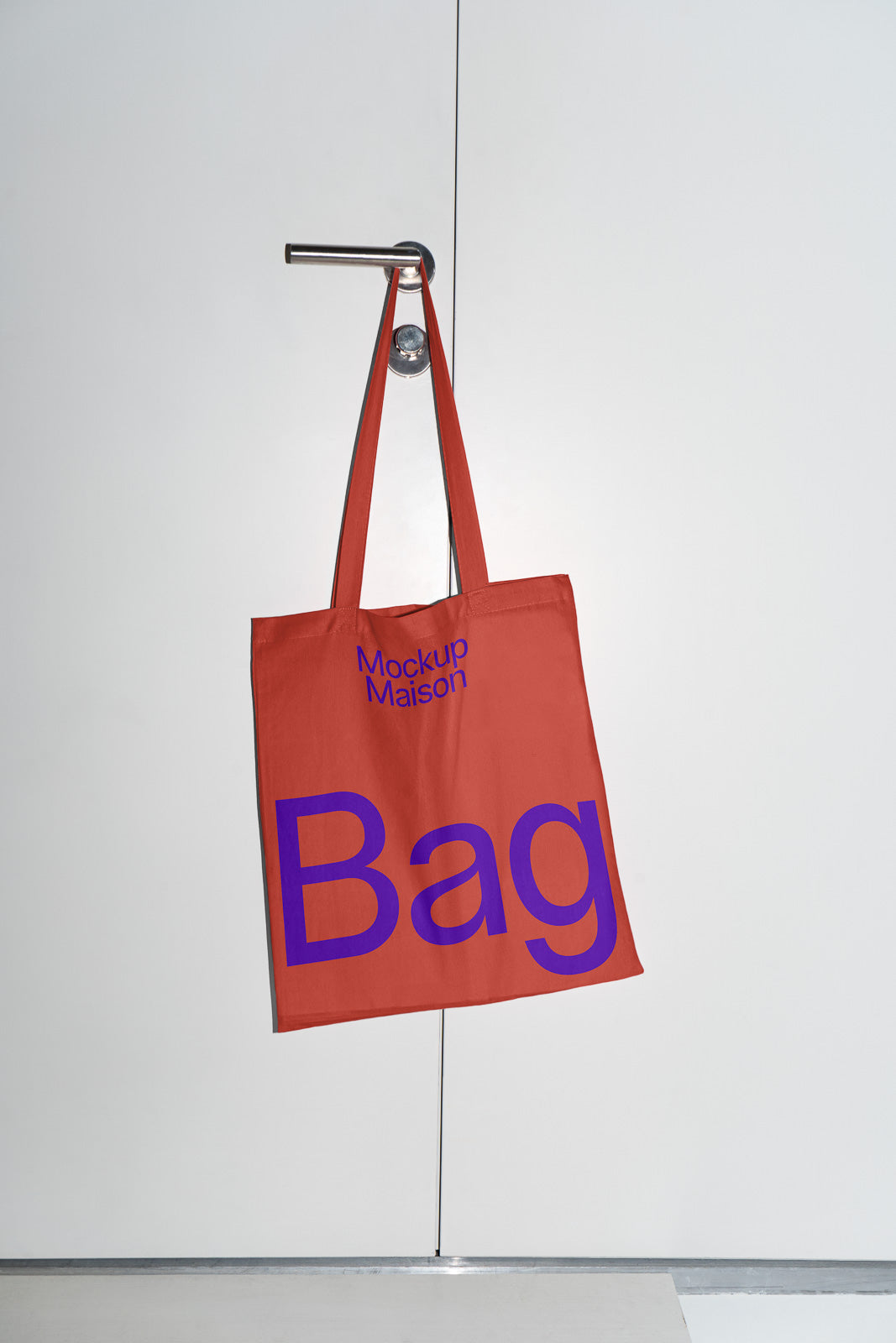 PSD mockup of a red tote bag with the words "Mockup Maison" and "Bag" printed in large purple letters, hanging on a metal door handle.