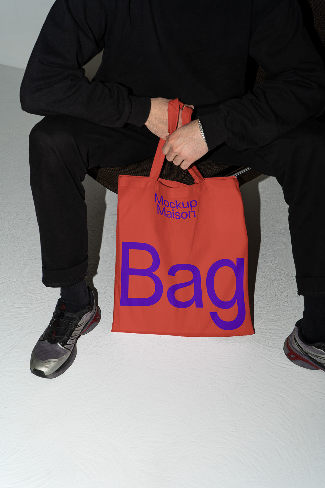 PSD mockup of a person holding an orange tote bag with blue text reading "Mockup Maison Bag," seated against a neutral background.