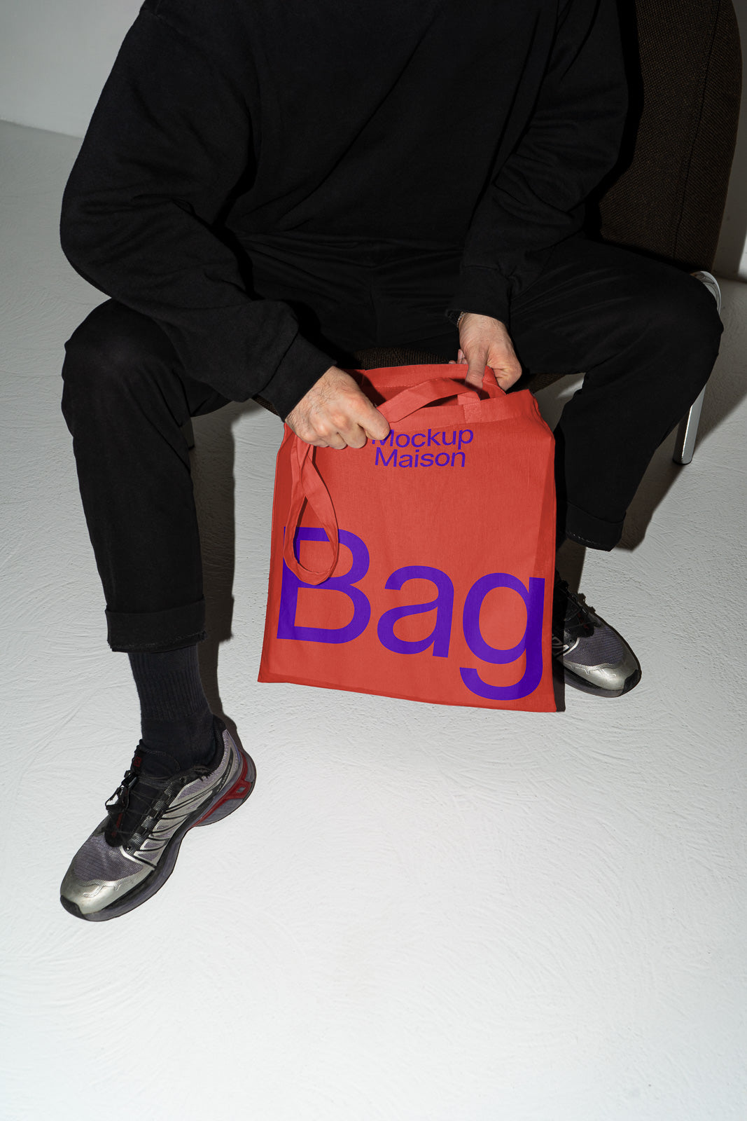 PSD mockup of a person holding a red tote bag with large blue text design, sitting on a chair in a minimalist setting.