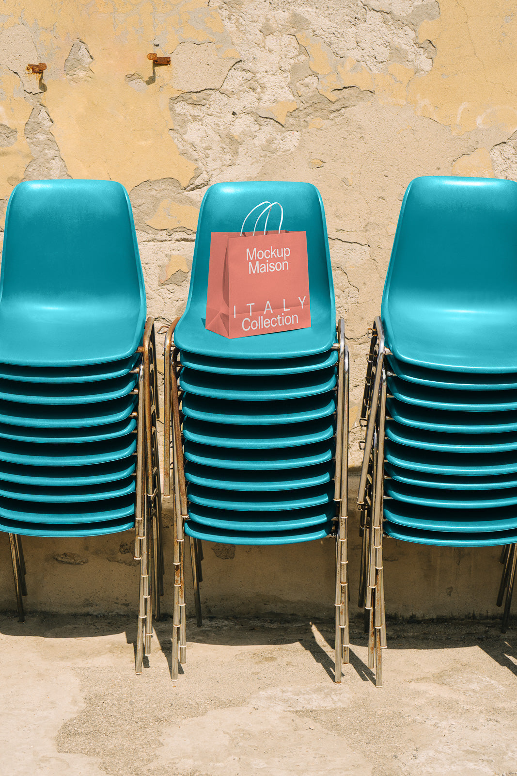 PSD mockup of a pink paper bag labeled "Mockup Maison Italy Collection" placed on a stack of teal chairs against a textured wall.