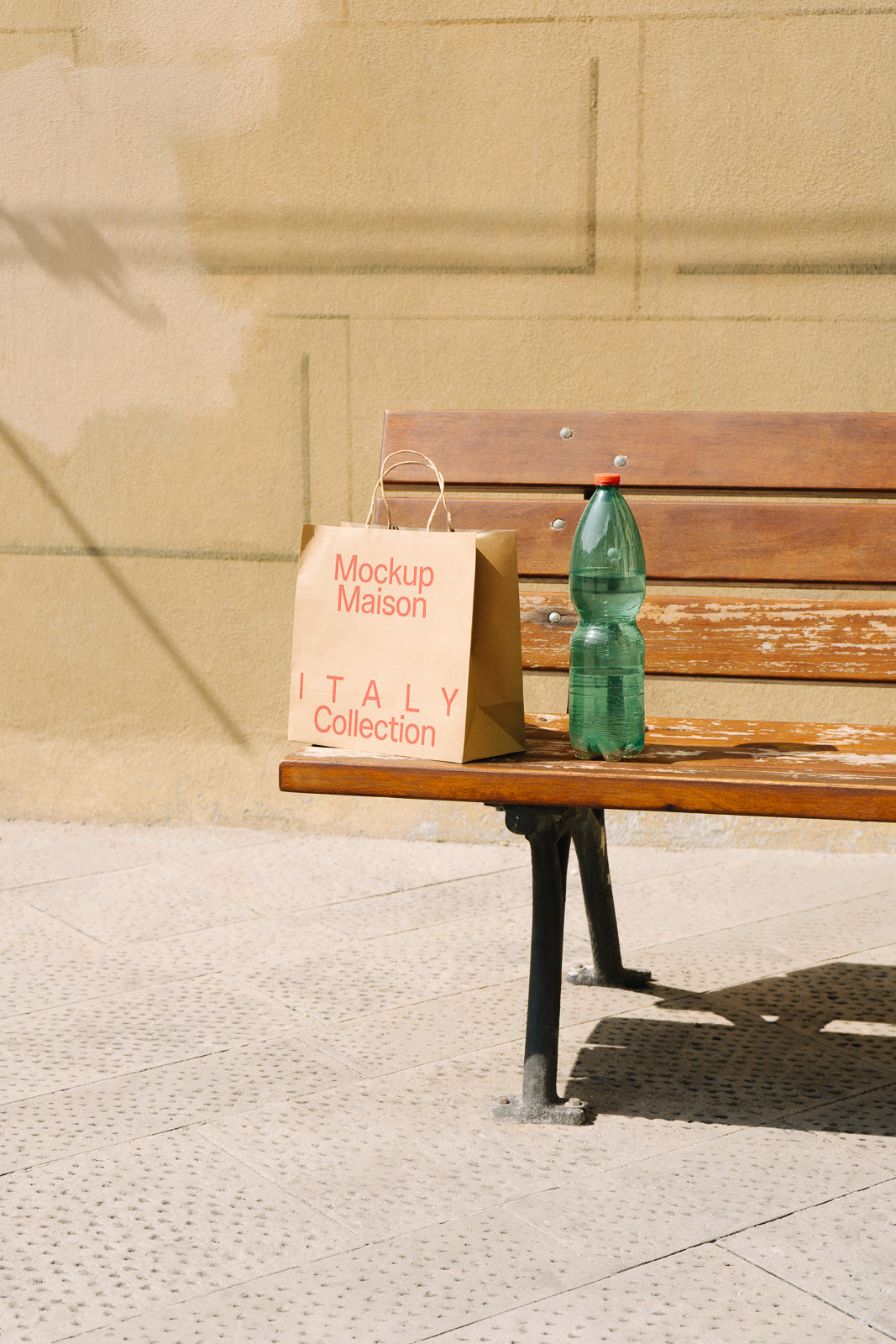 PSD mockup of a paper bag labeled "Mockup Maison Italy Collection" beside a green plastic bottle on a wooden bench against a textured wall background.
