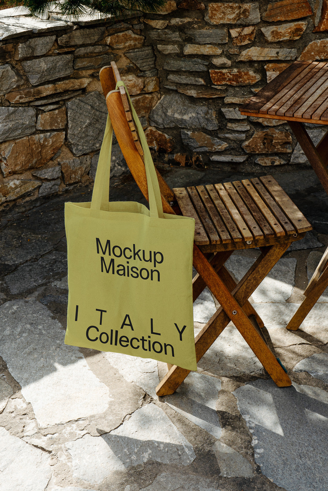 PSD mockup of a green tote bag hanging on a wooden chair with text "Mockup Maison Italy Collection" against a stone wall background.
