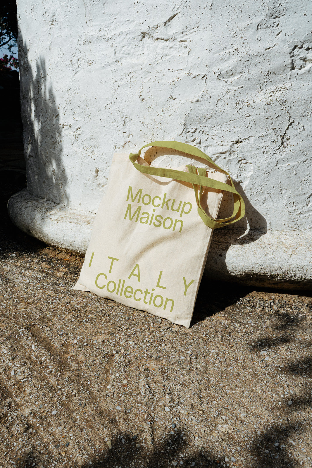 PSD mockup of a canvas tote bag labeled "Mockup Maison Italy Collection," leaning against a stone structure on a textured ground.