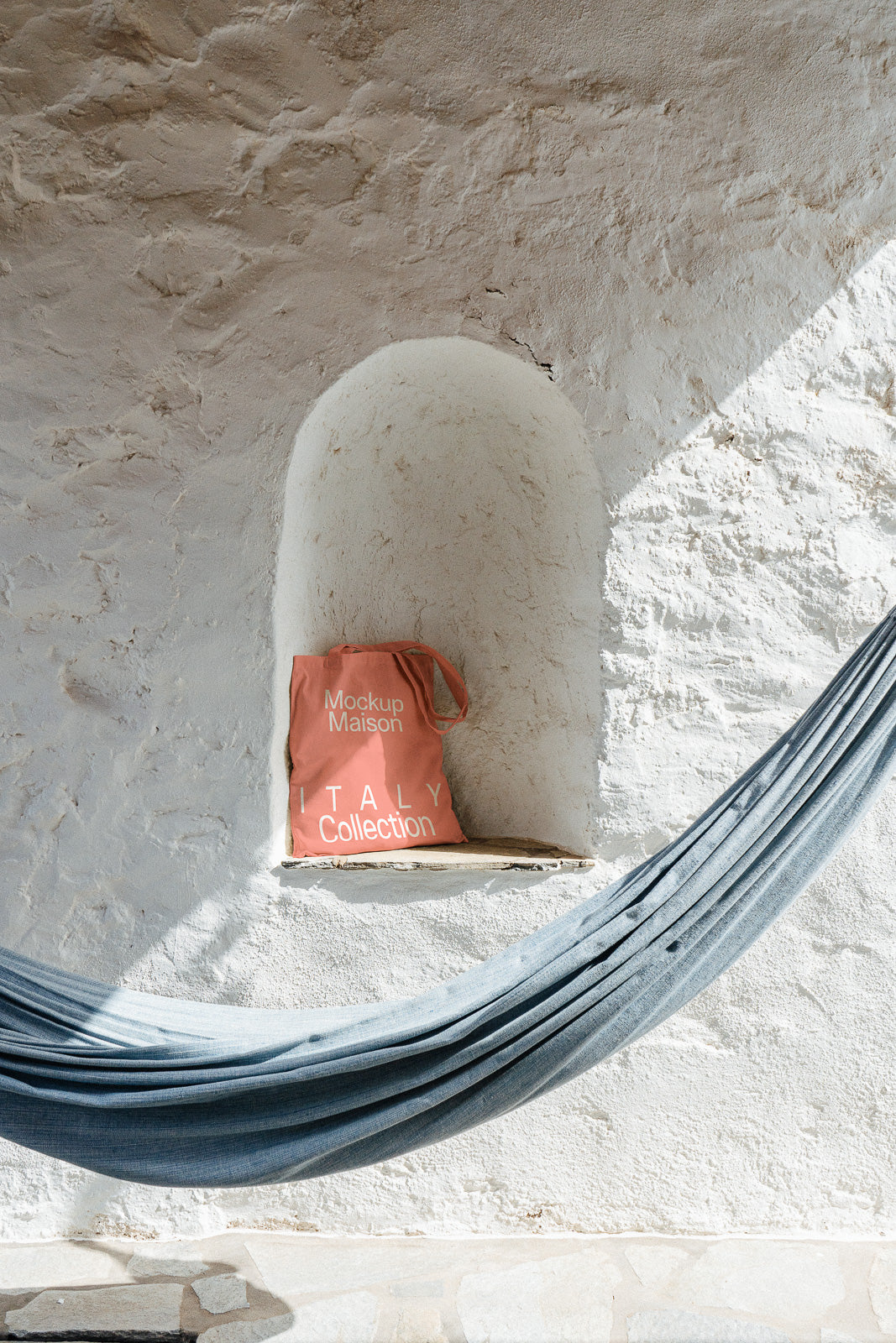PSD mockup of a coral tote bag with "Mockup Maison ITALY Collection" text on a stone shelf, next to a draped hammock against a white textured wall.