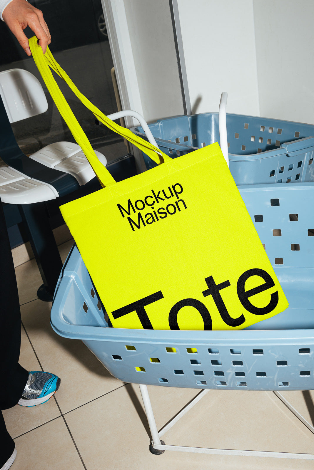 PSD mockup of a bright yellow tote bag with bold black text, placed in a blue laundry basket indoors, with part of a shoe visible nearby.