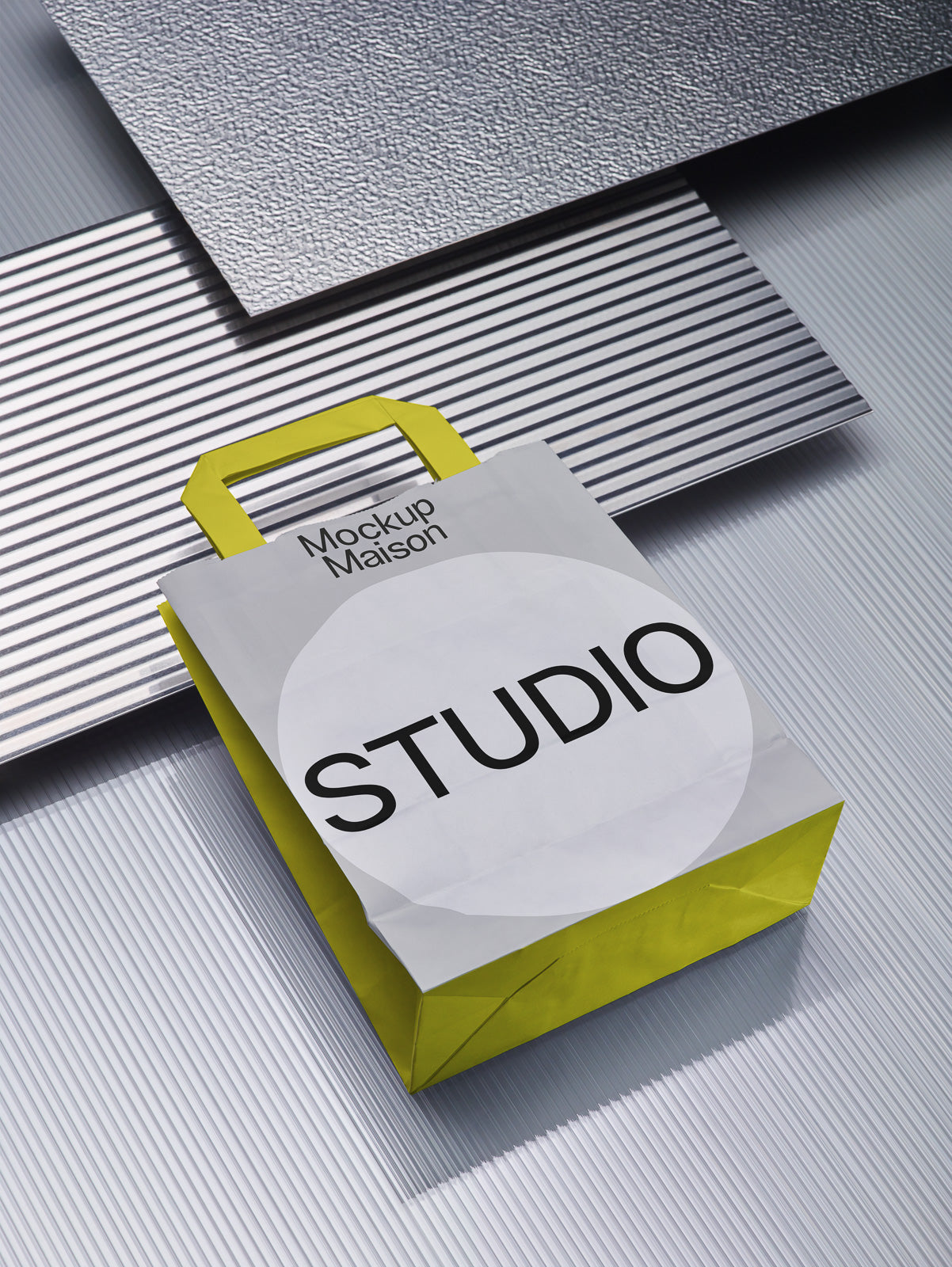 PSD mockup of a white paper bag with yellow sides and handles, showing "Mockup Maison STUDIO" text, placed on a textured, metallic surface.