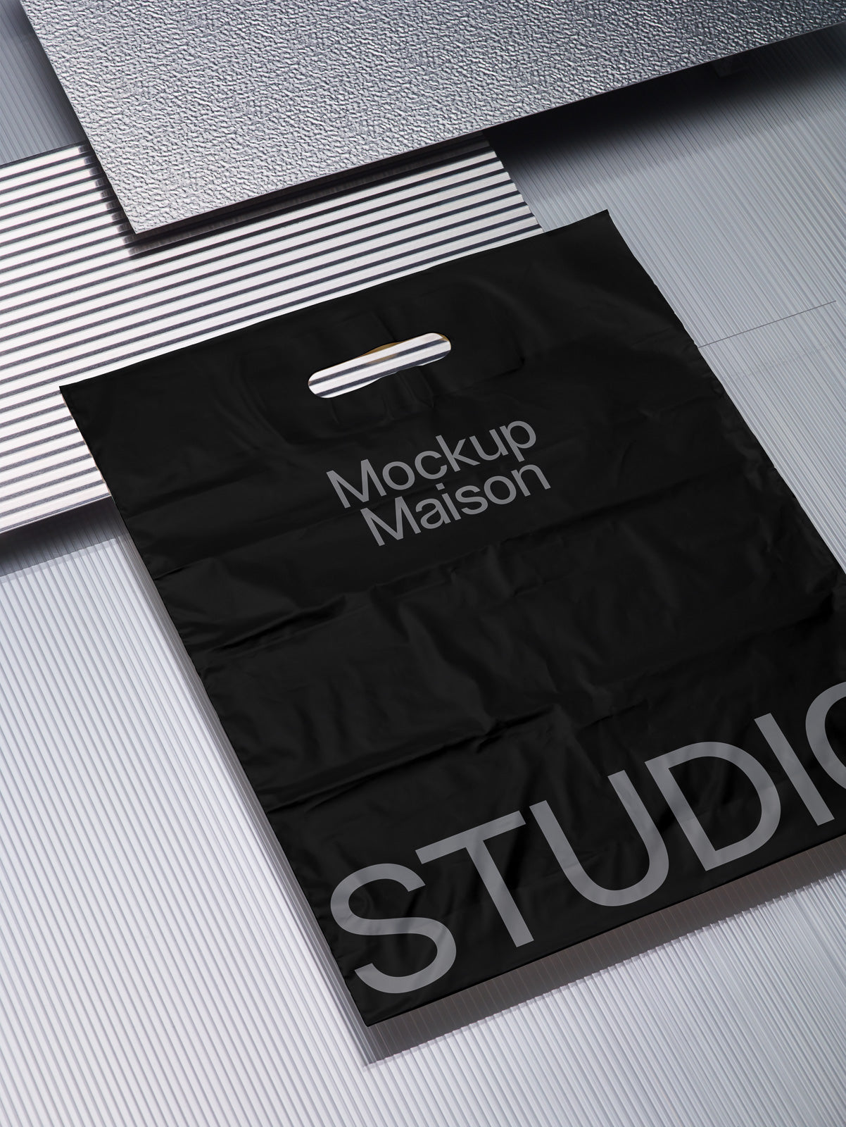 PSD mockup of a black plastic bag with "Mockup Maison" and "STUDIO" text, placed on a textured metallic and striped background.
