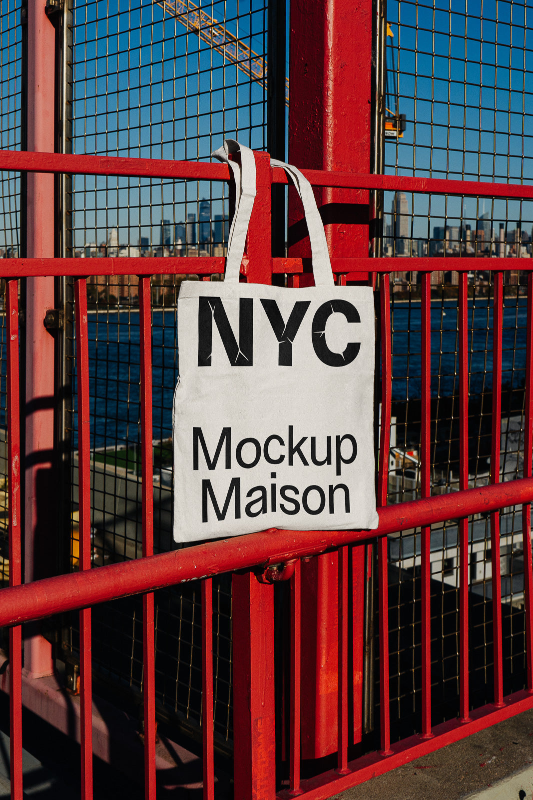 PSD mockup of a white tote bag with the text "NYC Mockup Maison" hanging on a red metal fence with an urban skyline in the background.