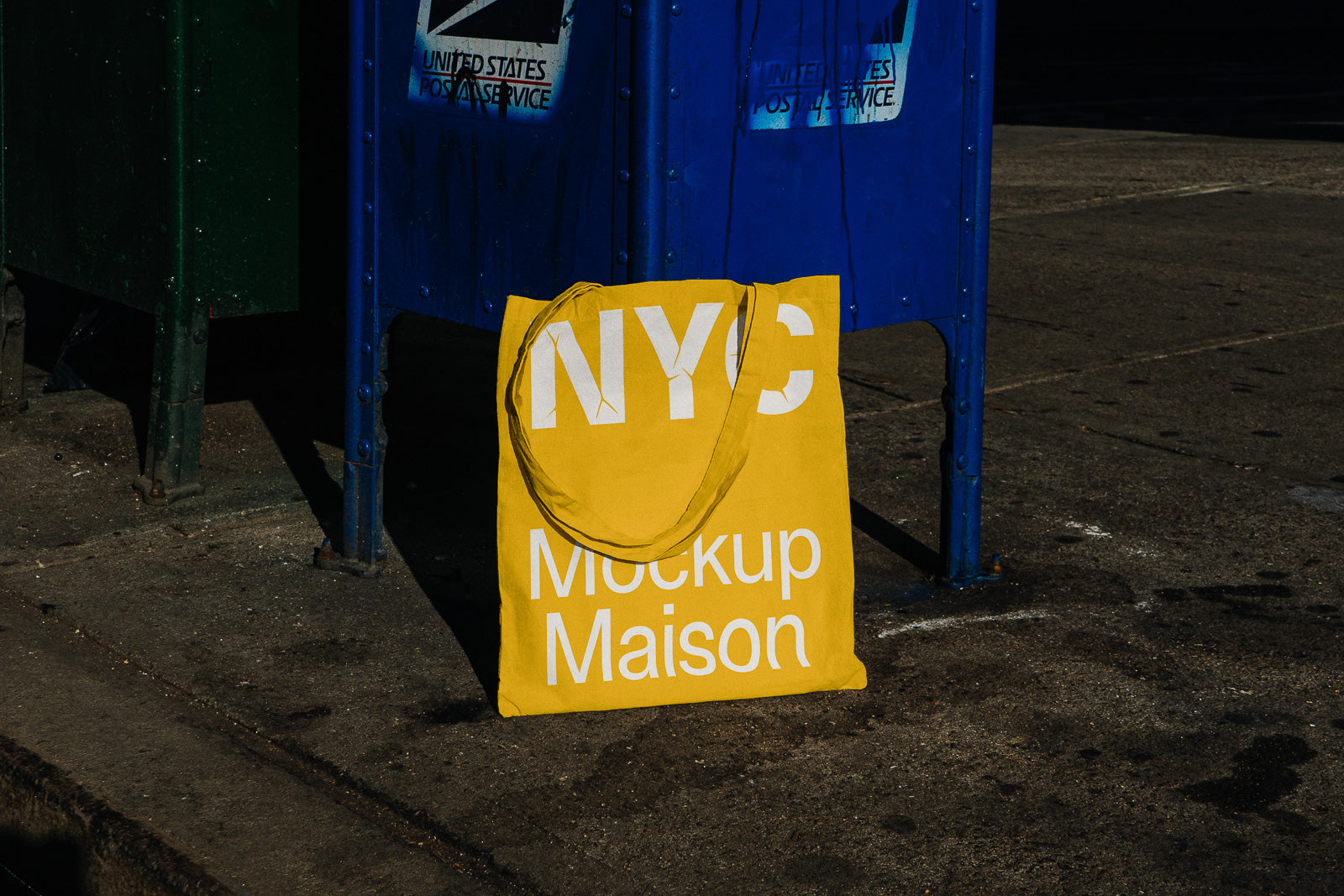 PSD mockup of a yellow tote bag with "NYC Mockup Maison" text, leaning against a blue mailbox on an urban sidewalk.