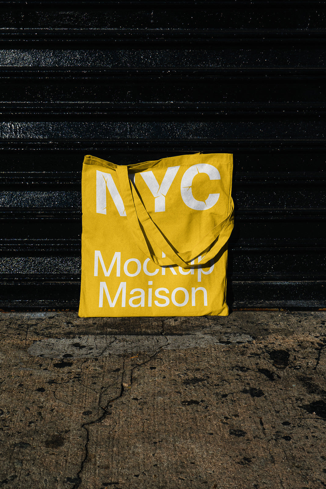 PSD mockup of a yellow tote bag with "NYC Mockup Maison" in white lettering, placed against a dark textured wall on concrete ground.