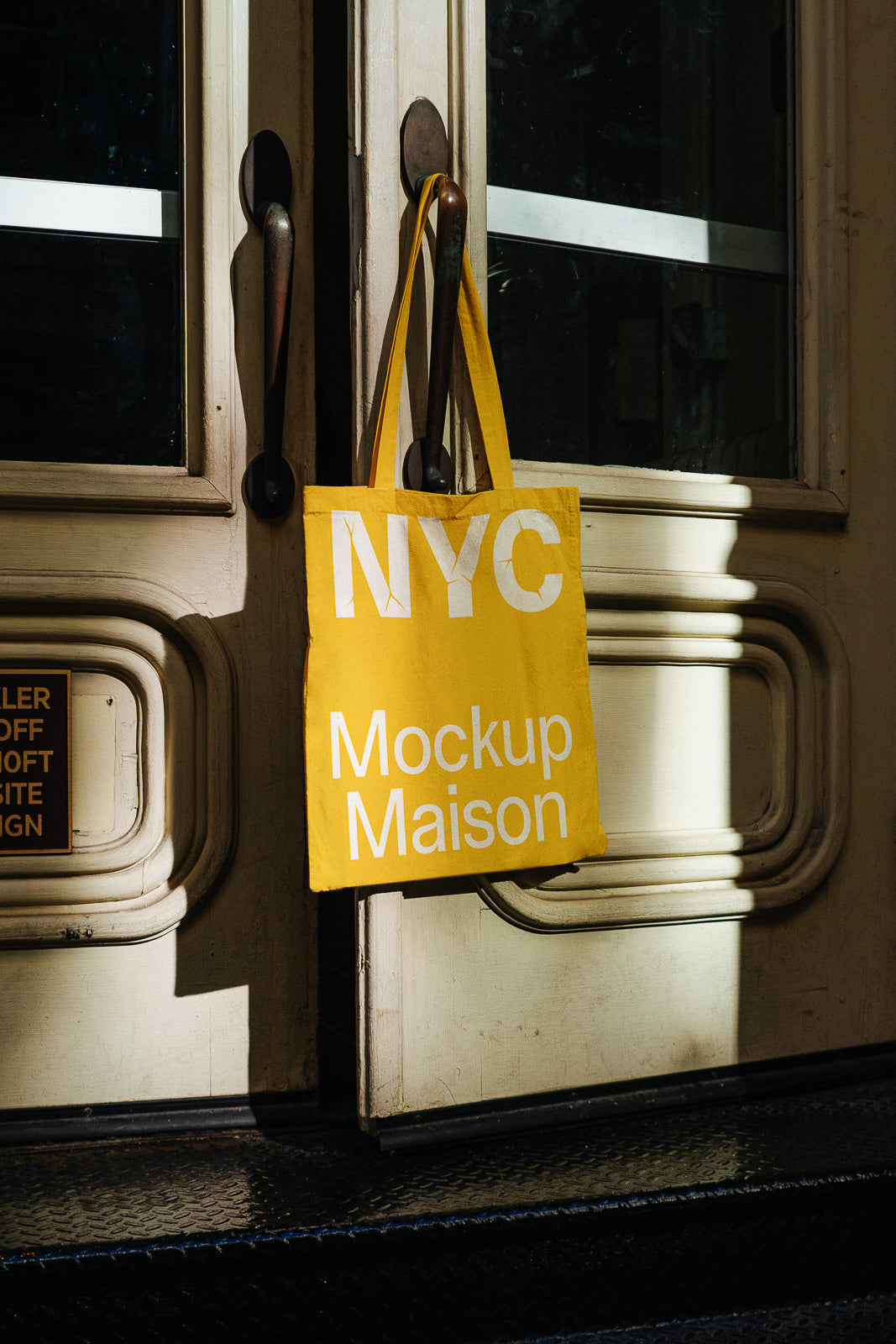 PSD mockup of a yellow tote bag with the text "NYC Mockup Maison" hanging on a vintage door handle, casting shadows on the wooden surface.