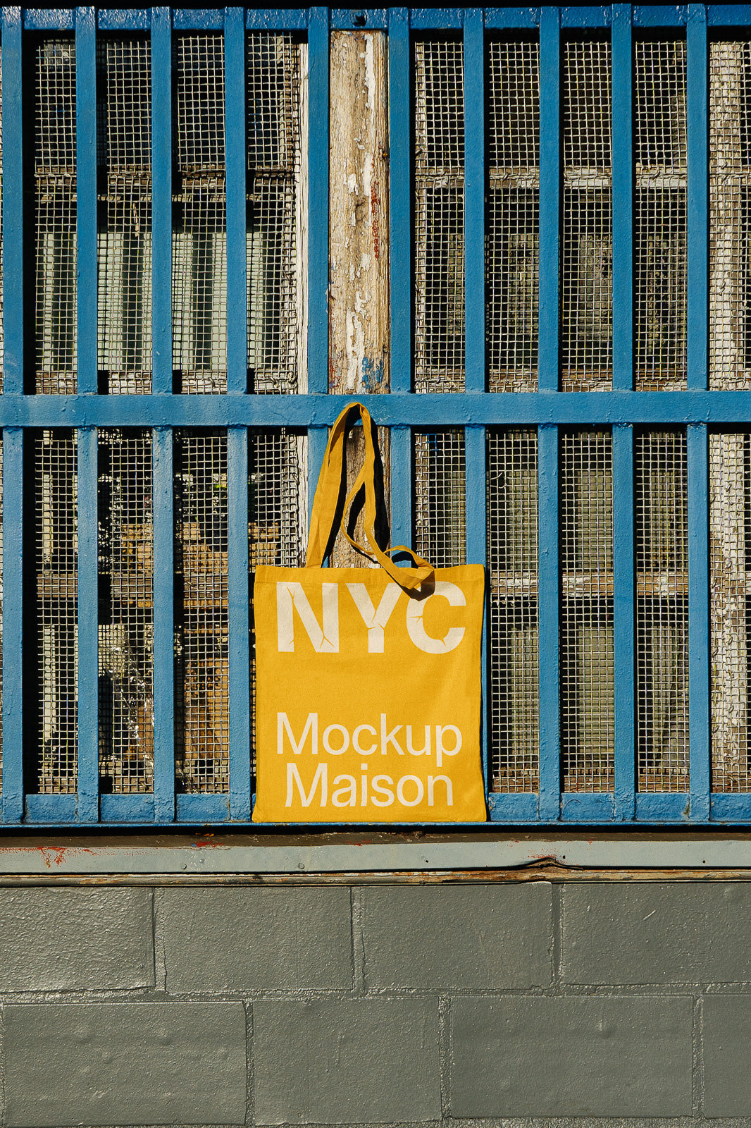 PSD mockup of a yellow tote bag labeled "NYC Mockup Maison," hanging on a textured blue metal fence with mesh panels in an urban setting.
