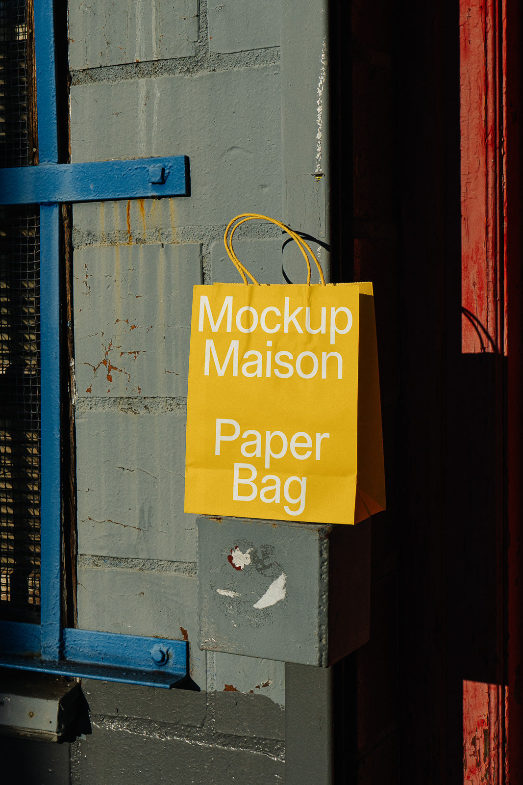 PSD mockup of a yellow paper bag with text "Mockup Maison Paper Bag" leaning against a textured gray wall and blue metal frame.