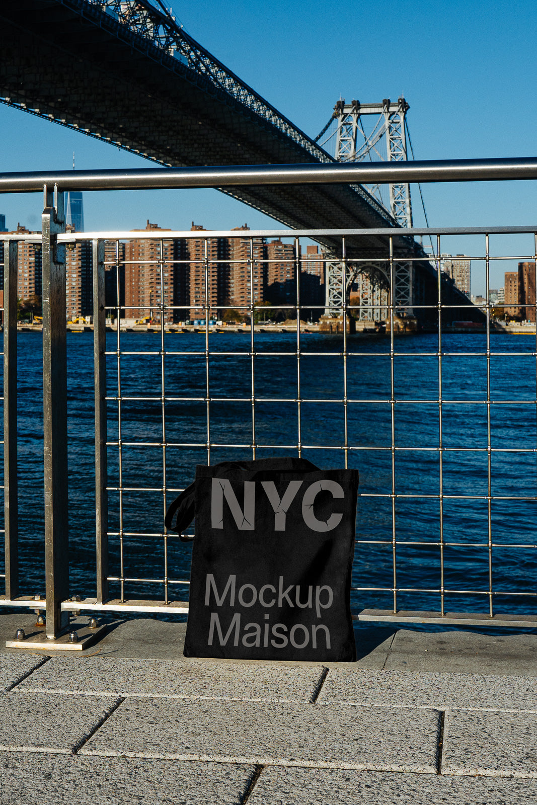 PSD mockup of a black tote bag with "NYC Mockup Maison" text, situated by a waterfront with a bridge and cityscape in the background.