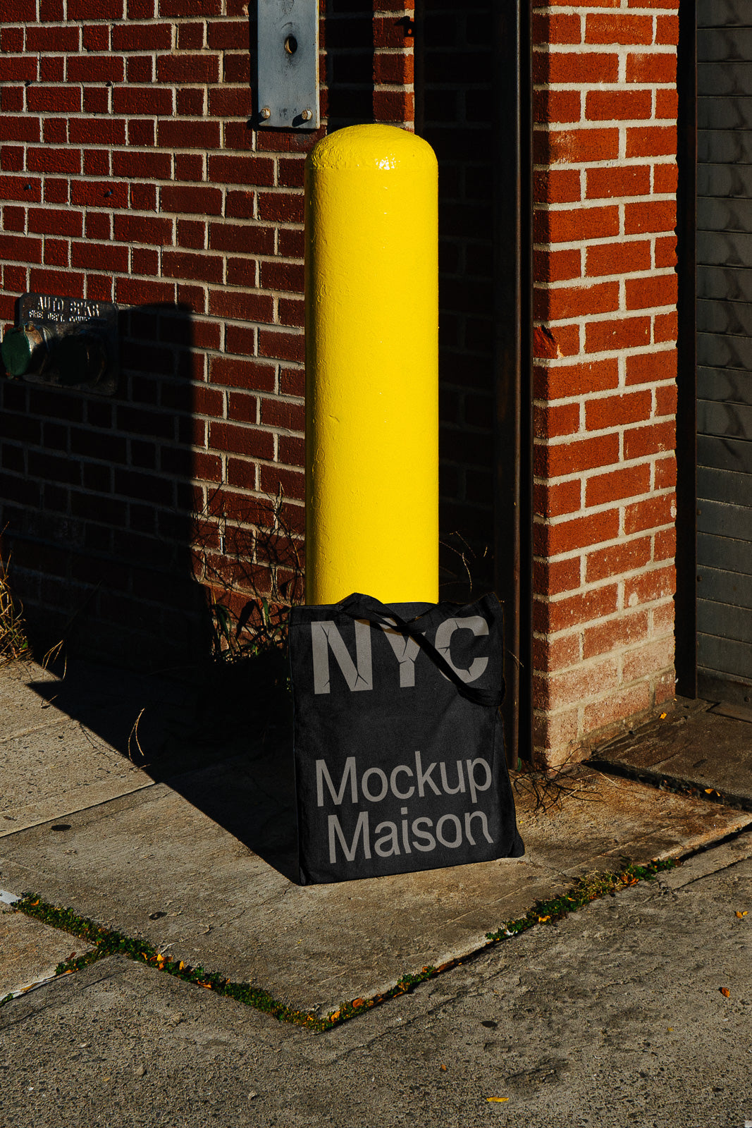 PSD mockup of a black tote bag leaning against a yellow bollard and brick wall, featuring the text "NYC Mockup Maison" in a street setting.