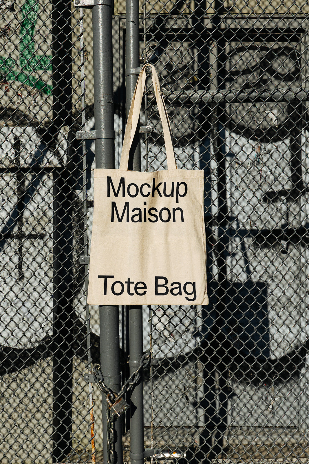 PSD mockup of a tote bag hanging on a chain-link fence, displaying the text "Mockup Maison Tote Bag" in bold letters, with a padlock below.