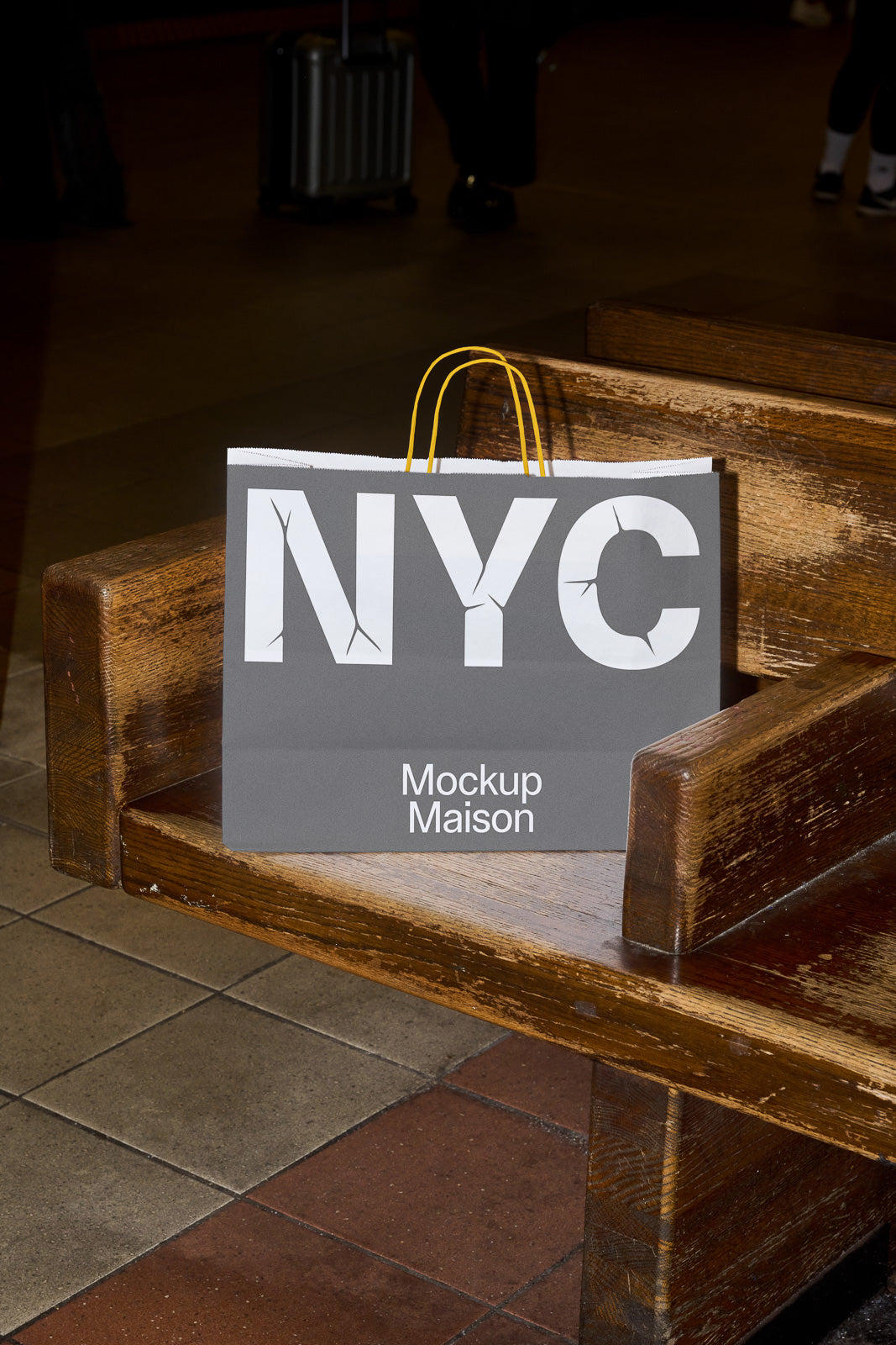 PSD mockup of a paper shopping bag with "NYC" and "Mockup Maison" text, placed on a wooden bench in a public setting.