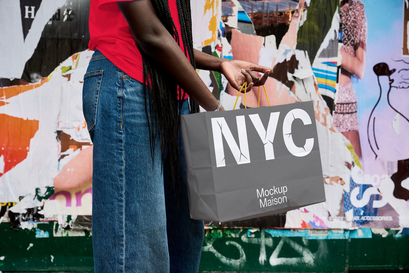 PSD mockup of a person holding a gray shopping bag with "NYC" text against a colorful, urban poster-covered wall background.