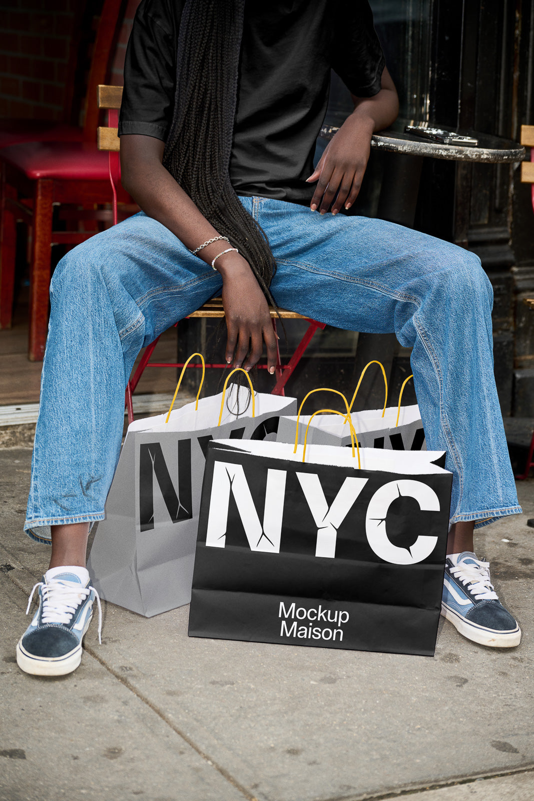 PSD mockup of NYC-themed shopping bags with bold lettering and yellow handles, placed on a sidewalk next to a person in jeans and sneakers.