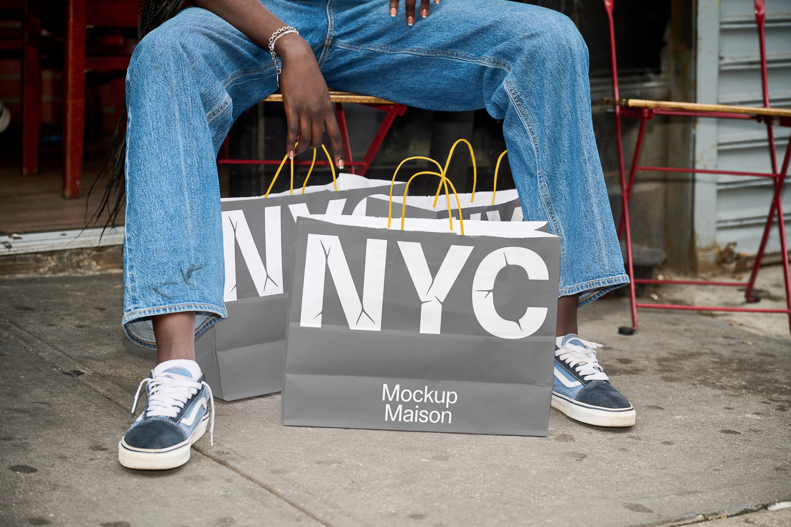PSD mockup of a person sitting outside holding multiple shopping bags with "NYC" and "Mockup Maison" printed, dressed in casual denim and sneakers.