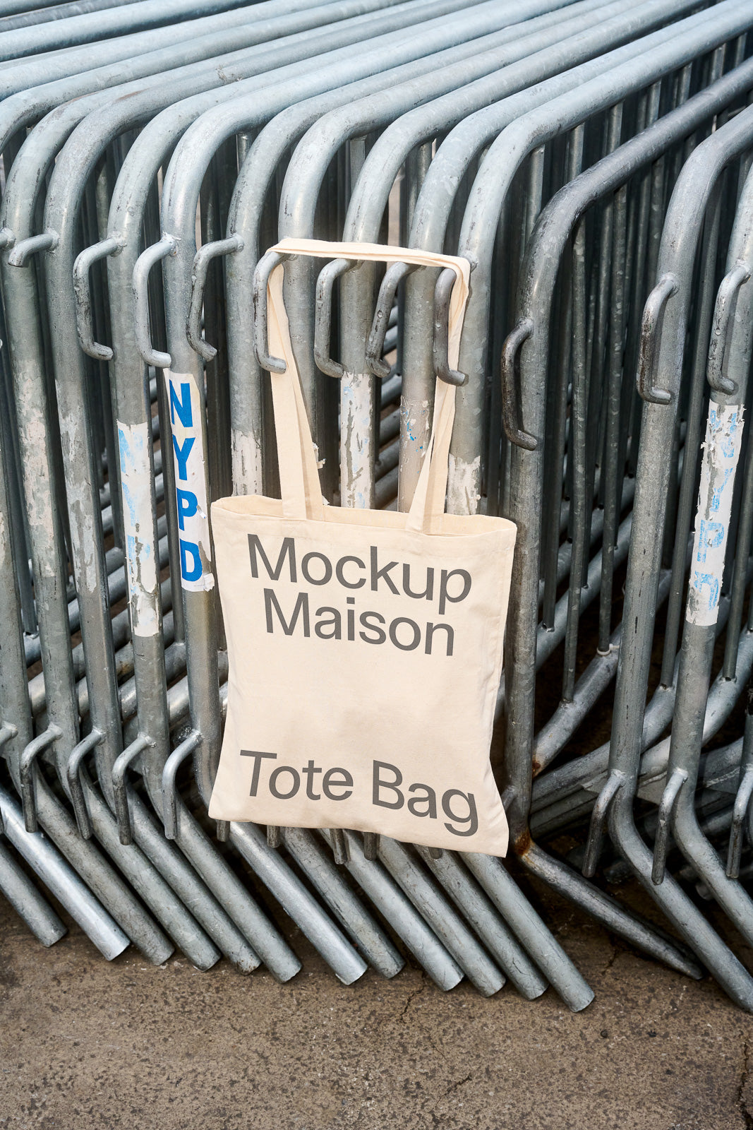 PSD mockup of a canvas tote bag with "Mockup Maison Tote Bag" printed on it, hanging on metal barricades.