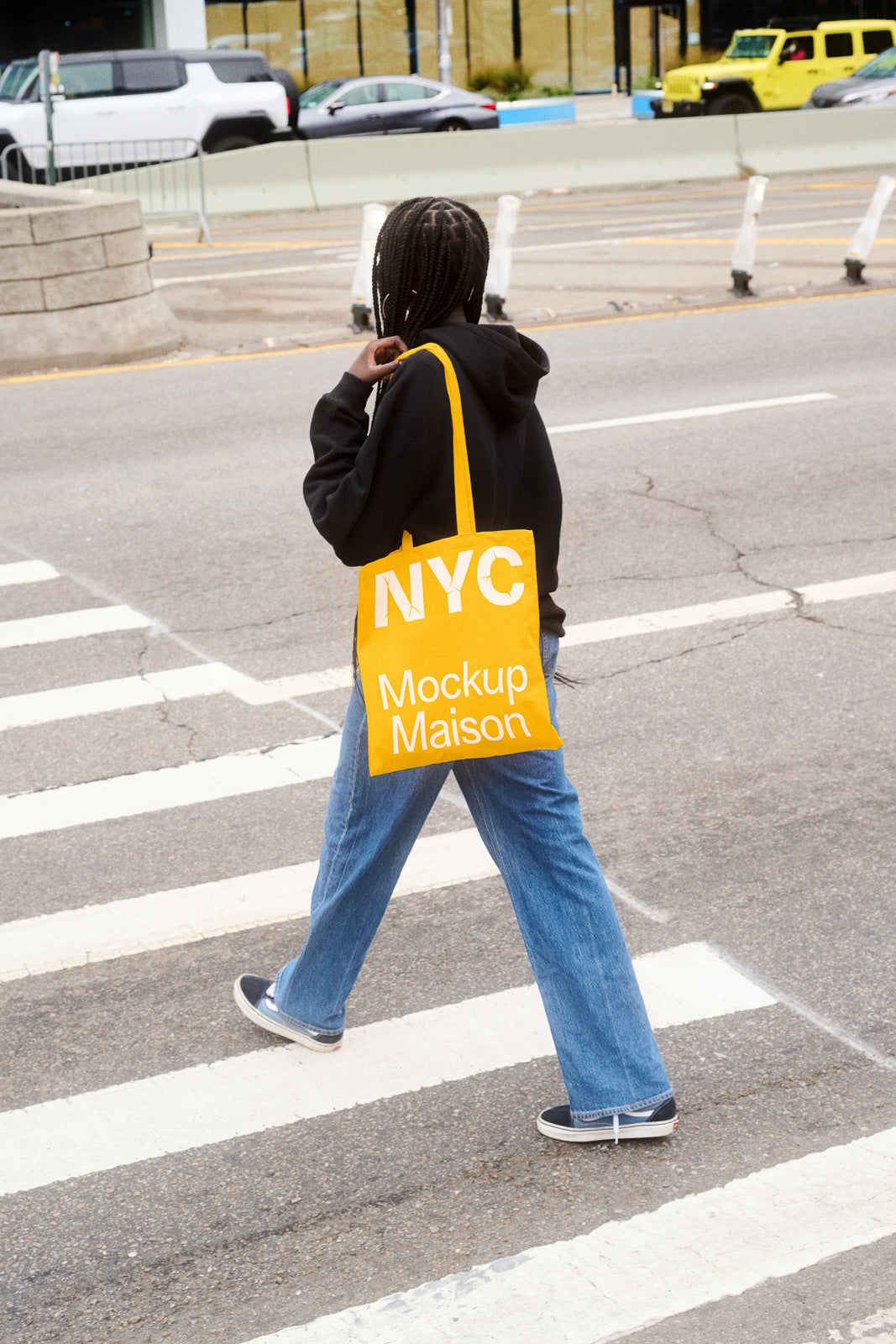 PSD mockup of a person crossing the street with a yellow tote bag labeled "NYC Mockup Maison," wearing a black hoodie and blue jeans.