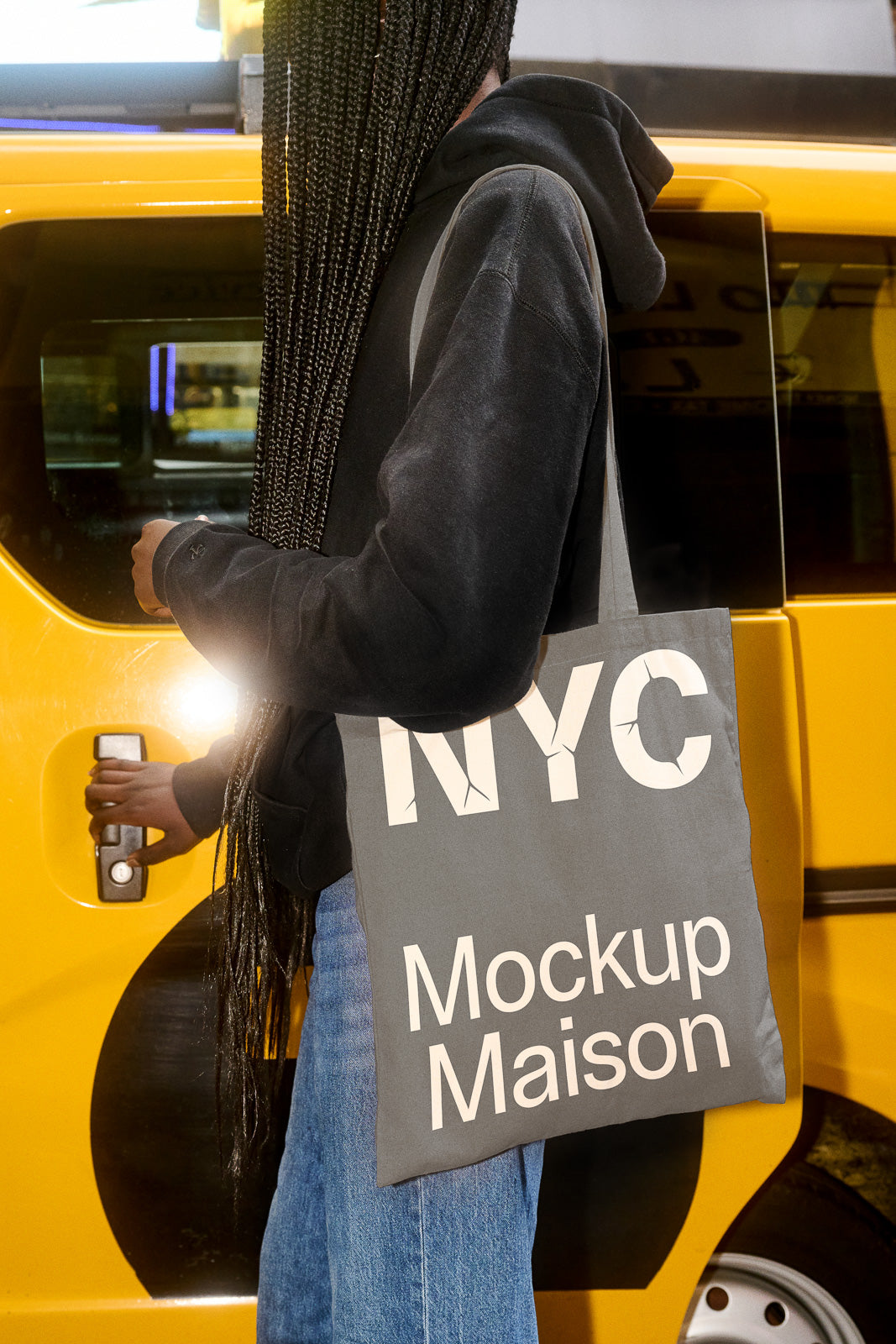 PSD mockup of a person holding a tote bag labeled "NYC Mockup Maison" against the background of a yellow taxi.