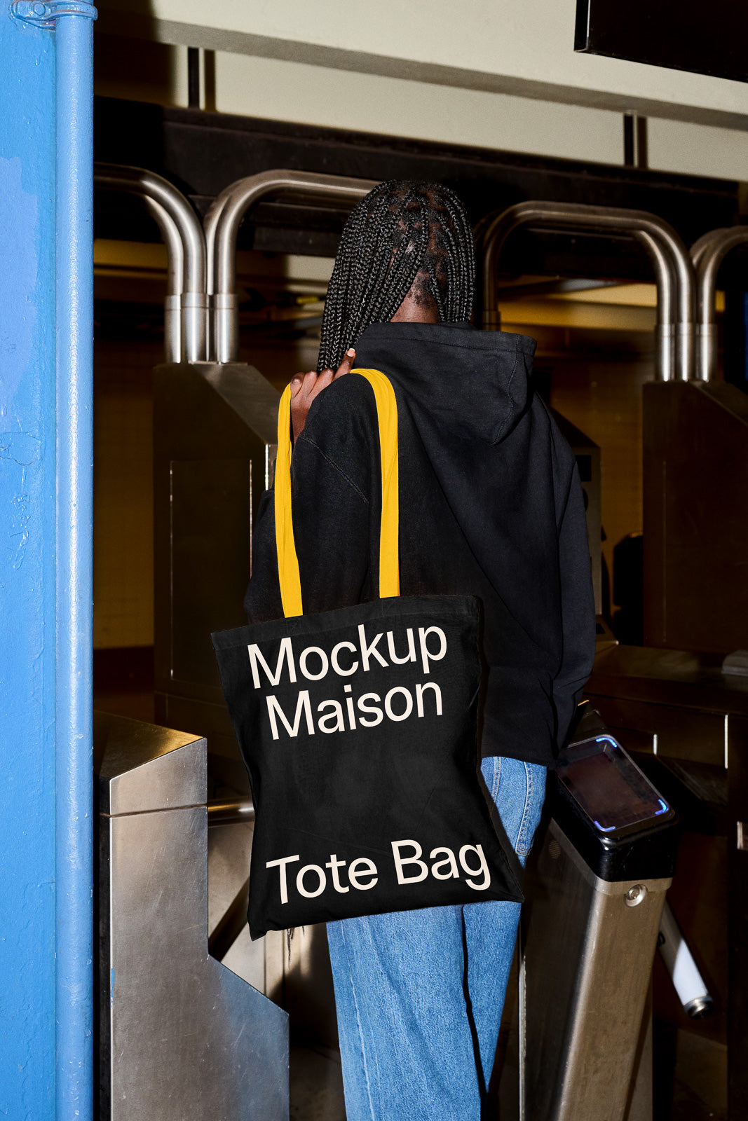 PSD mockup of a person with a black tote bag featuring "Mockup Maison Tote Bag" text, standing near a turnstile in a subway station.