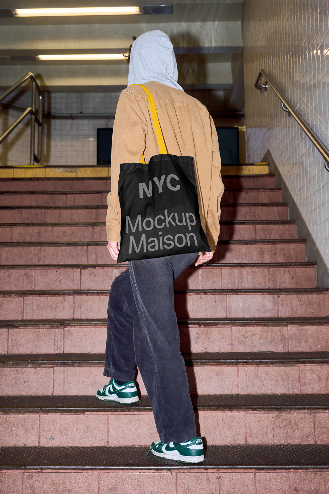 PSD mockup of a person in a hoodie and pants walking up stairs with a black tote bag labeled "NYC Mockup Maison" in a subway station.