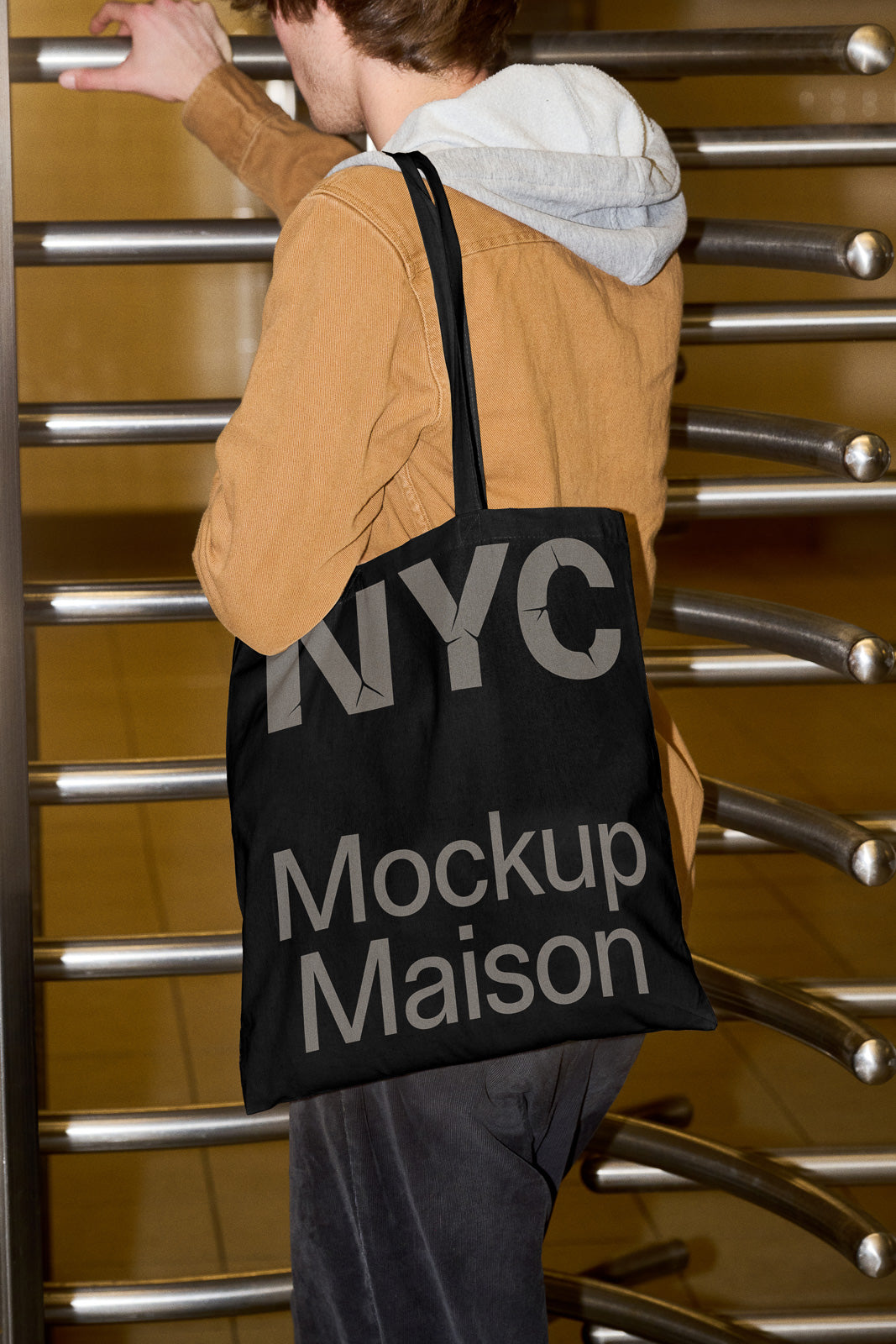 PSD mockup of a person in a brown jacket holding a black tote bag with "NYC Mockup Maison" text, standing by a metallic turnstile.
