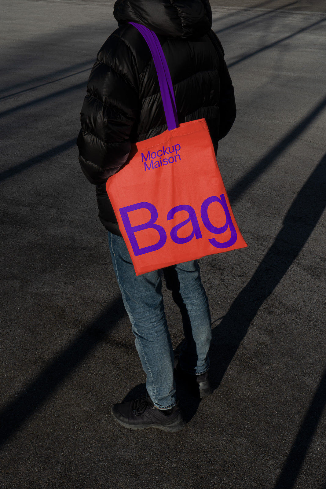 PSD mockup of a person wearing a black jacket and jeans, holding an orange tote bag with purple lettering reading "Mockup Maison Bag" in a shadowed outdoor setting.