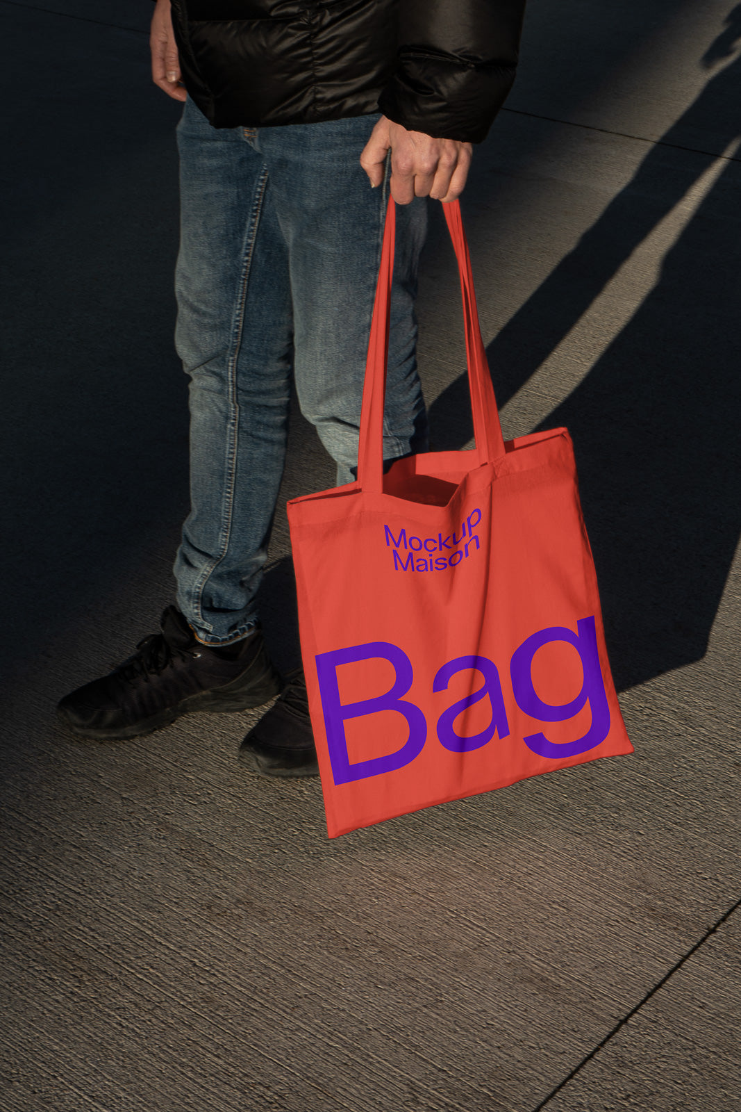 PSD mockup of a person holding a red tote bag with blue text reading "Mockup Maison Bag," shown in a shadowed outdoor setting.