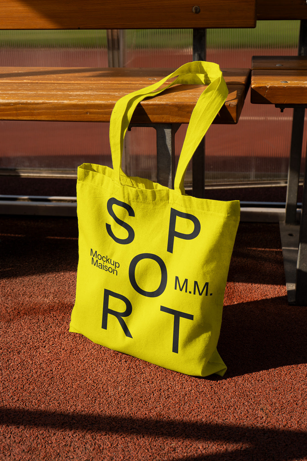 PSD mockup of a bright yellow tote bag with bold black letters spelling "SPORT," placed on the ground near a wooden bench in an outdoor setting.