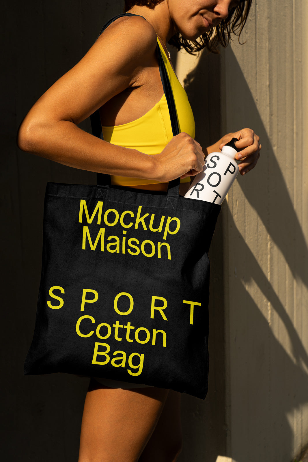 PSD mockup of a person holding a black cotton bag with "Mockup Maison SPORT" in yellow text, and a water bottle labeled "SPORT."
