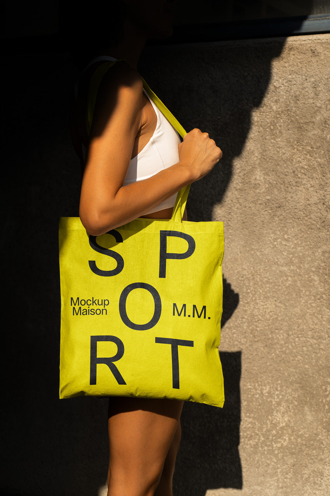 PSD mockup of a person holding a bright yellow tote bag with bold black lettering spelling "SPORT" and smaller text saying "Mockup Maison M.M."