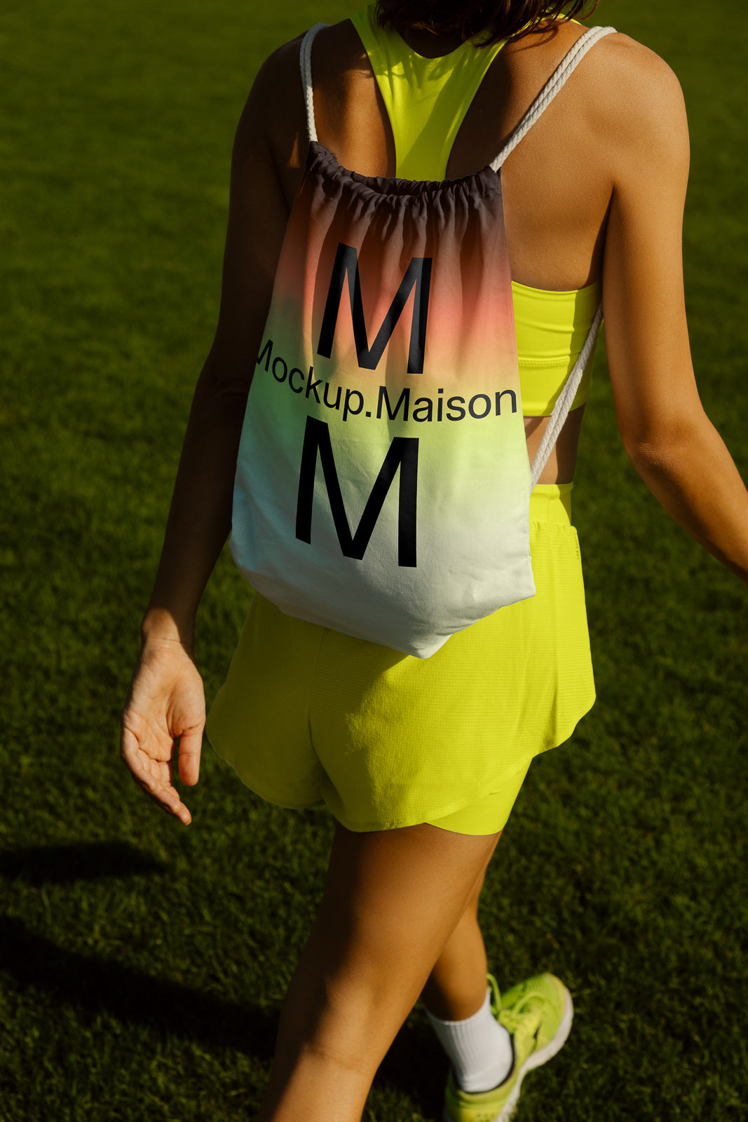PSD mockup of a drawstring backpack with "Mockup.Maison" text, worn by a person in neon activewear walking on grass.