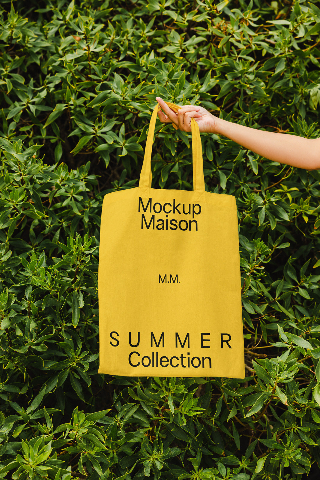 PSD mockup of a yellow tote bag with "Mockup Maison" and "Summer Collection" text, held by a hand, with a background of lush green foliage.