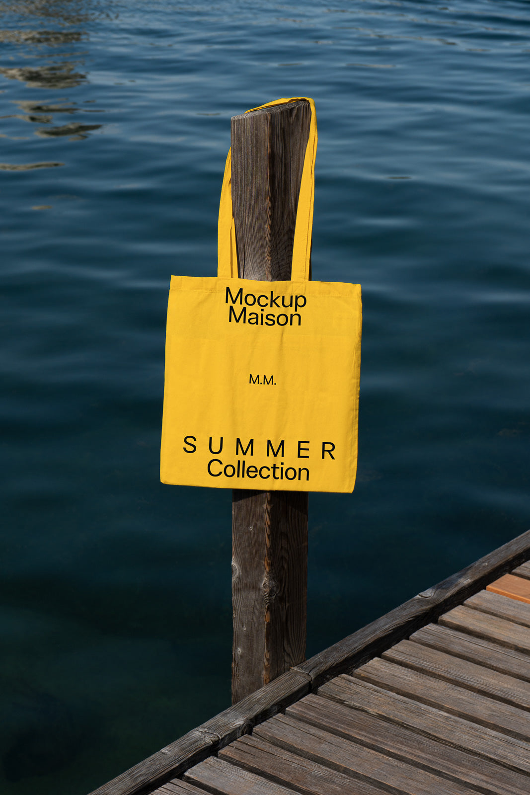 PSD mockup of a yellow tote bag hanging from a wooden post by a waterfront, featuring "Mockup Maison" and "Summer Collection" text.