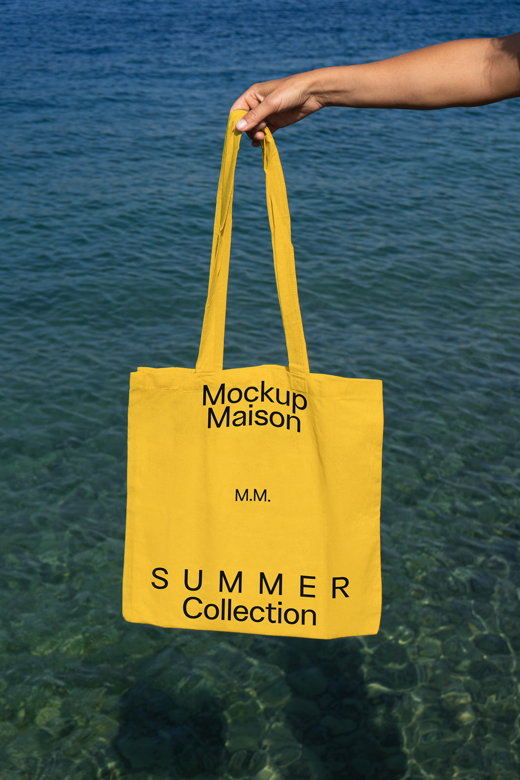 PSD mockup of a yellow tote bag with "Mockup Maison" and "Summer Collection" text, held by a person above clear blue water.