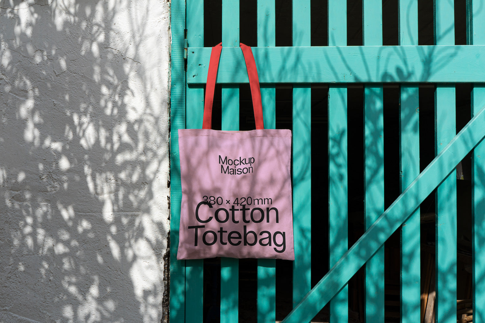 PSD mockup of a pink cotton tote bag with red handles hanging on a teal gate, showcasing the text "Mockup Maison 380 x 420 mm Cotton Totebag."
