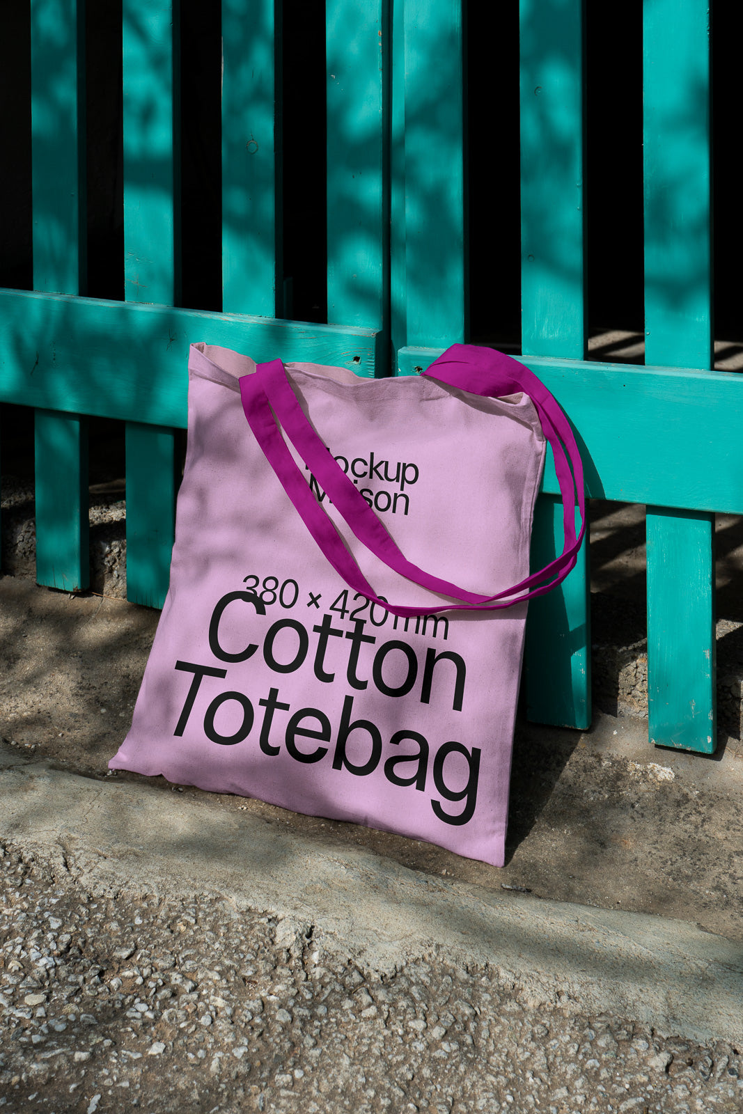 PSD mockup of a pink cotton tote bag with purple handles leaning against a blue wooden fence on a sunny day. Text on bag indicates size 380 x 420mm.