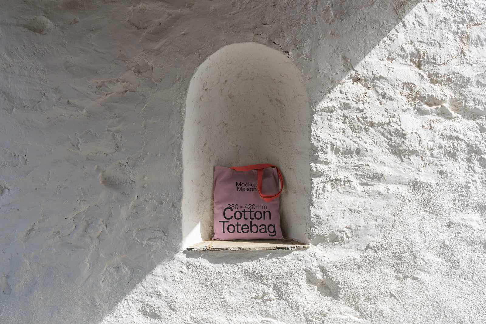 PSD mockup of a pink cotton tote bag with red straps, positioned in a recessed stone wall, showcasing its texture and design details.