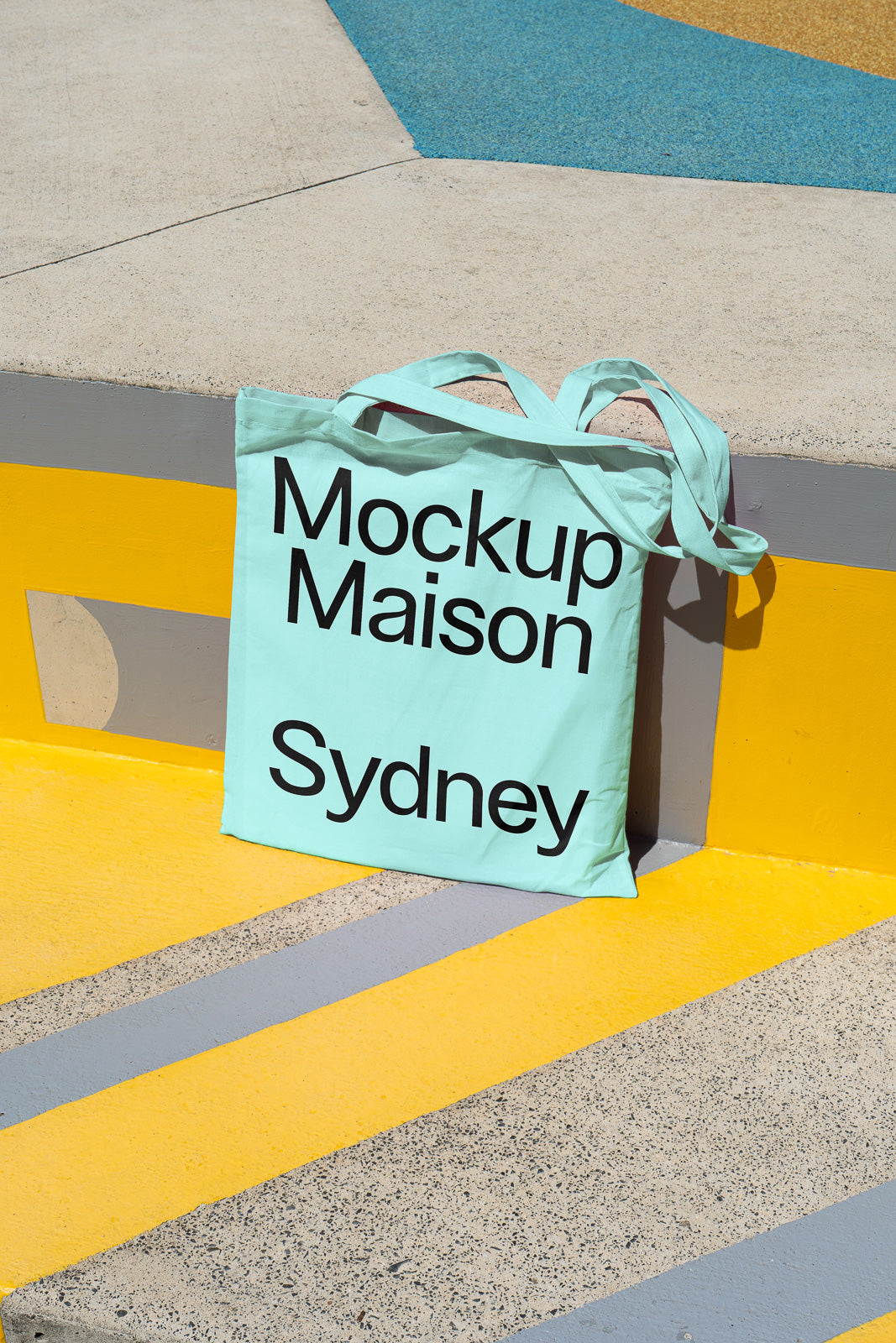 PSD mockup of a light blue tote bag with "Mockup Maison Sydney" text, placed on a colorful geometric background with yellow and gray accents.