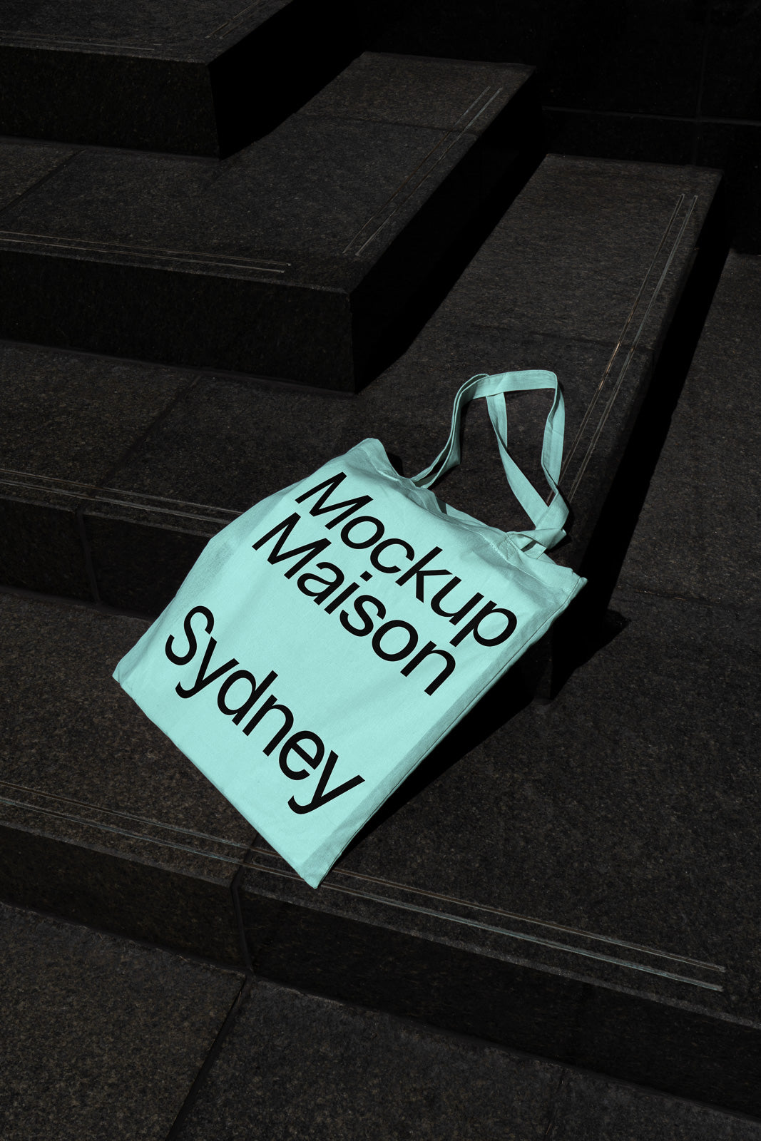 PSD mockup of a light blue tote bag placed on dark stone steps, with bold black text reading "Mockup Maison Sydney."