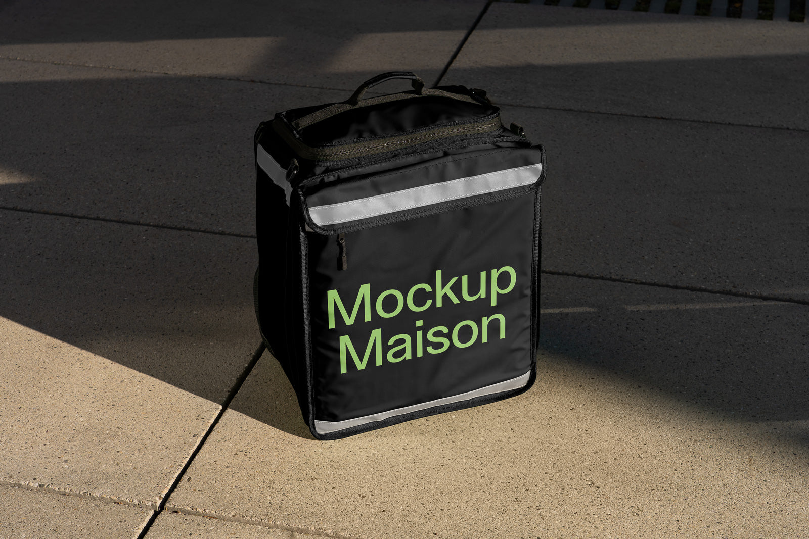 PSD mockup of a black delivery bag with reflective strips and "Mockup Maison" in green text on a sunlit concrete surface.