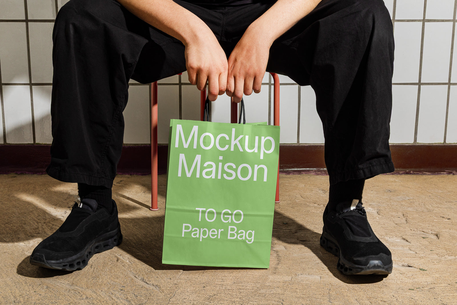 PSD mockup of a person sitting on a chair holding a green paper bag labeled "Mockup Maison TO GO Paper Bag," with a tiled wall backdrop.