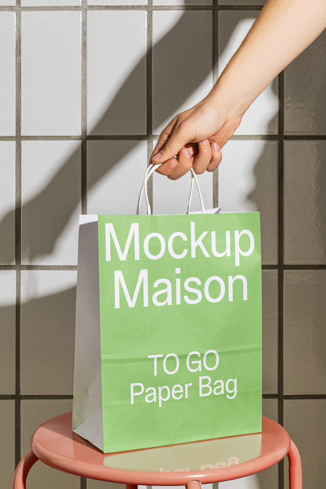 PSD mockup of a green paper bag labeled "Mockup Maison TO GO Paper Bag" being held by a hand. The bag rests on a pink stool against a tiled wall background.